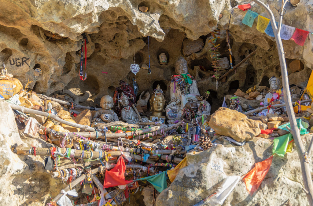 Santuario del Buda en Tarifa – Un altar espiritual en plena montaña de La Peña. Primer plano del Santuario del Buda en la Ruta del Buda de Tarifa, con esculturas de Buda, velas, piedras y amuletos espirituales colocados en una cueva natural en la roca. Detalle del Santuario del Buda en la Ruta del Buda de Tarifa. Se pueden ver varias figuras de Buda adornadas con amuletos, piedras y banderas tibetanas dentro de una cueva en la roca.🌿🕉️